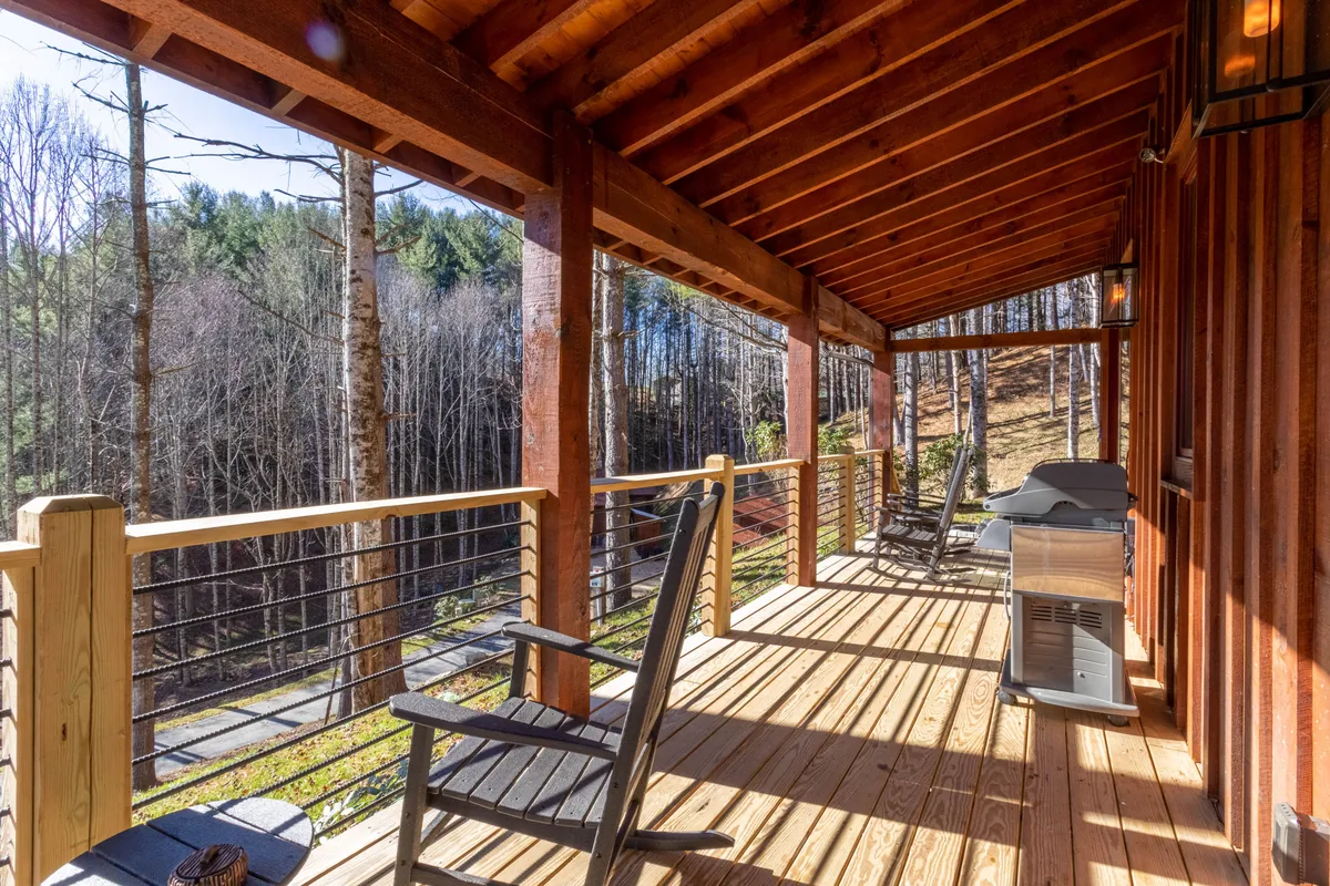 A tranquil, spacious wooden deck featuring inviting rocking chairs, a modern grill, and surrounded by lush trees. The setup offers a peaceful haven for relaxing, socializing, or preparing a barbecue while being immersed in nature.