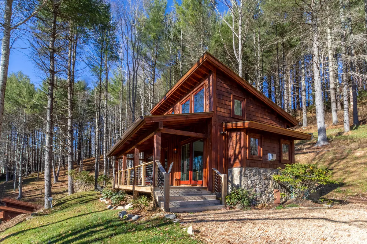 The image features a charming wooden cabin nestled among towering trees, with a welcoming front porch overlooking natural surroundings. The scene evokes tranquility, inviting guests to relax on the porch and enjoy sunsets or venture into the nearby forest trails.