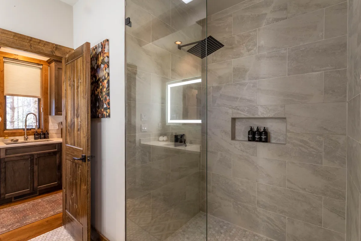 The bathroom shown highlights a modern, glass-enclosed curbless shower with a sleek rainfall showerhead, complemented by natural wood accents and a contemporary vanity with a dark cabinet and sink. The space is bright, clean, and evokes a spa-like serenity.