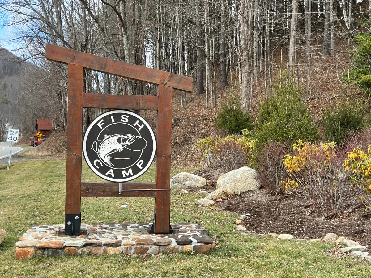 A close-up of a wooden sign structure adorned with a fish logo, surrounded by lush greenery. The image evokes the spirit of outdoor adventure, suggesting fishing spots and nature walks nearby.
