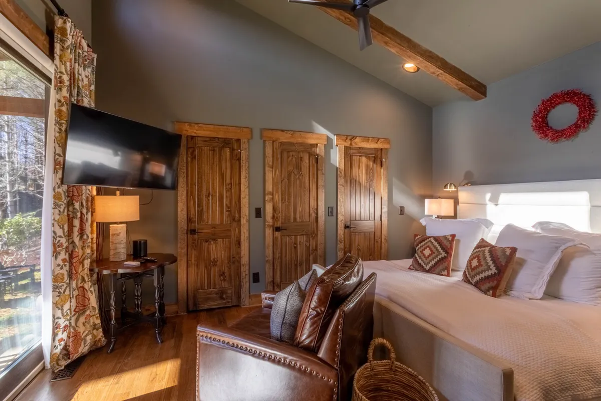 This image features a bedroom with a plush bed adorned with decorative pillows, wooden doors lending a warm rustic vibe, a smart TV visible on the wall, a table lamp casting gentle light, and a comfortable armchair in view. Natural light streams in, creating an inviting and restful ambiance.