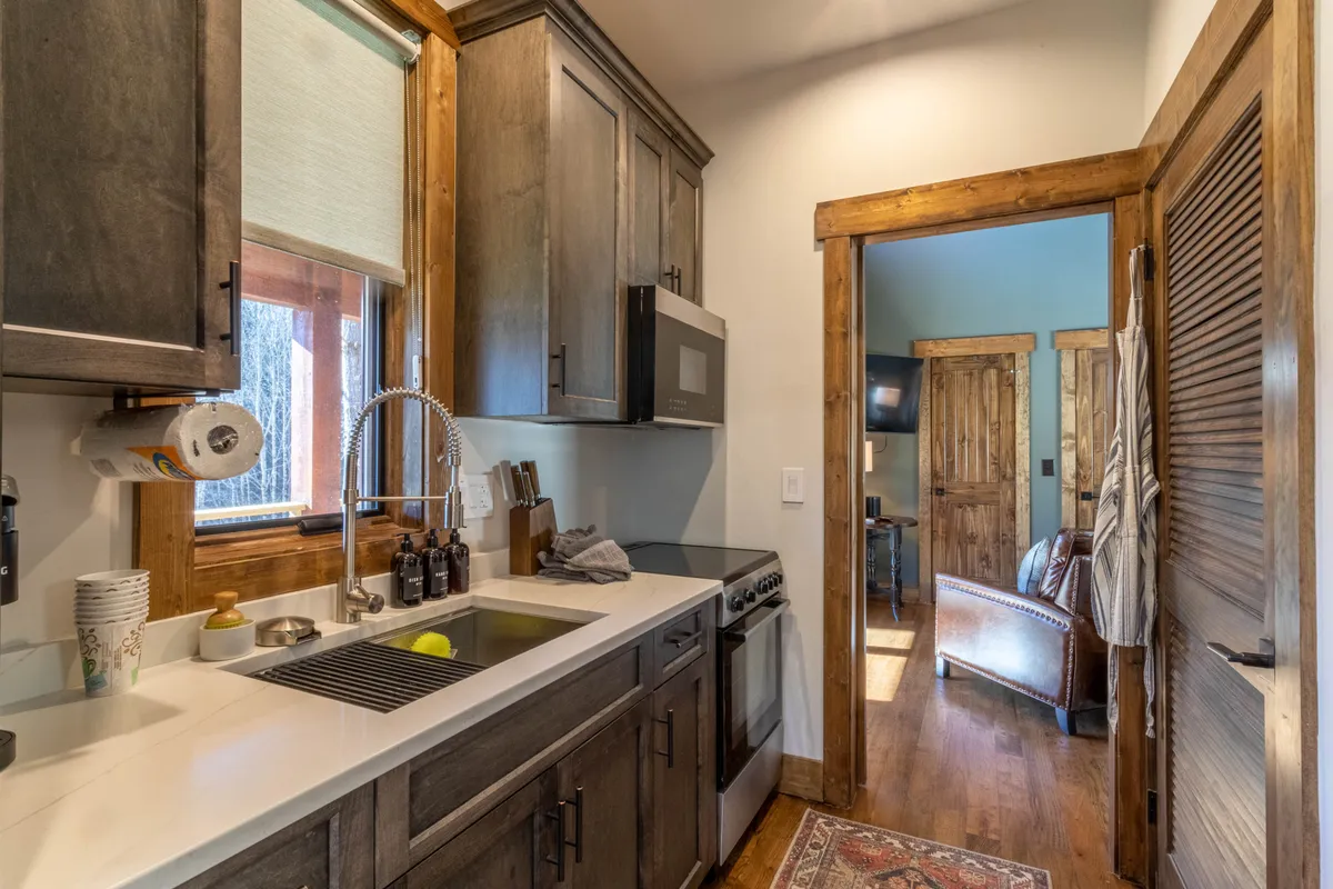 A modern interior view focused on a stylish kitchen sink with sleek fixtures, set against warm wooden cabinetry and soft recessed lighting. The open doorway offers a glimpse into the adjacent living area, evoking a sense of social flow and connection.