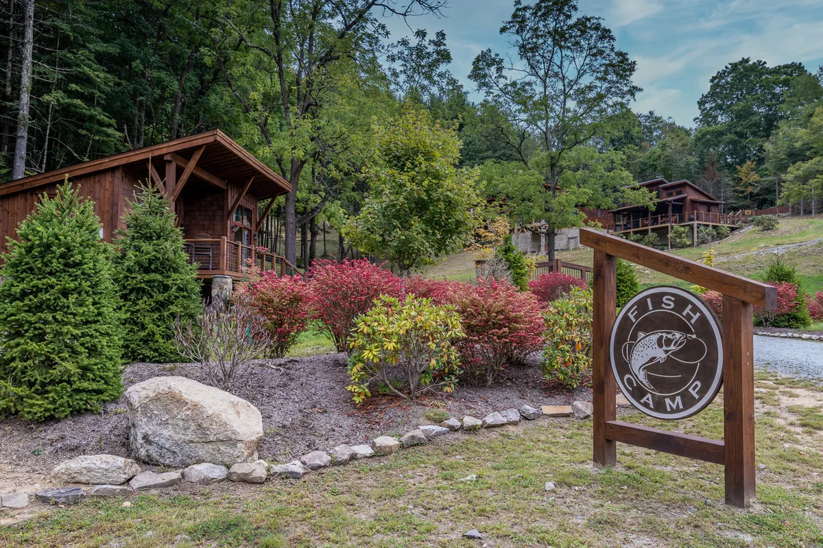 A collection of rustic wooden cabins is nestled amidst vibrant flower bushes and towering trees, with a signboard featuring a fish emblem. The setting suggests activities like fishing, hiking, and social gatherings, and radiates a relaxed, welcoming vibe.