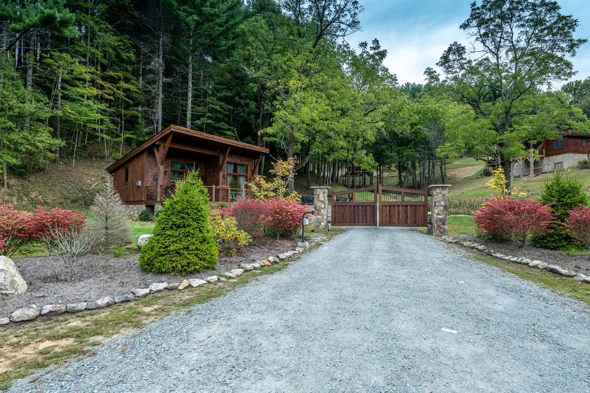 This image captures the exterior of a welcoming wooden cabin set within a landscaped garden and framed by a wooden gate and driveway. The scene is tranquil, with nature enveloping the home and inviting guests to enjoy the outdoors.