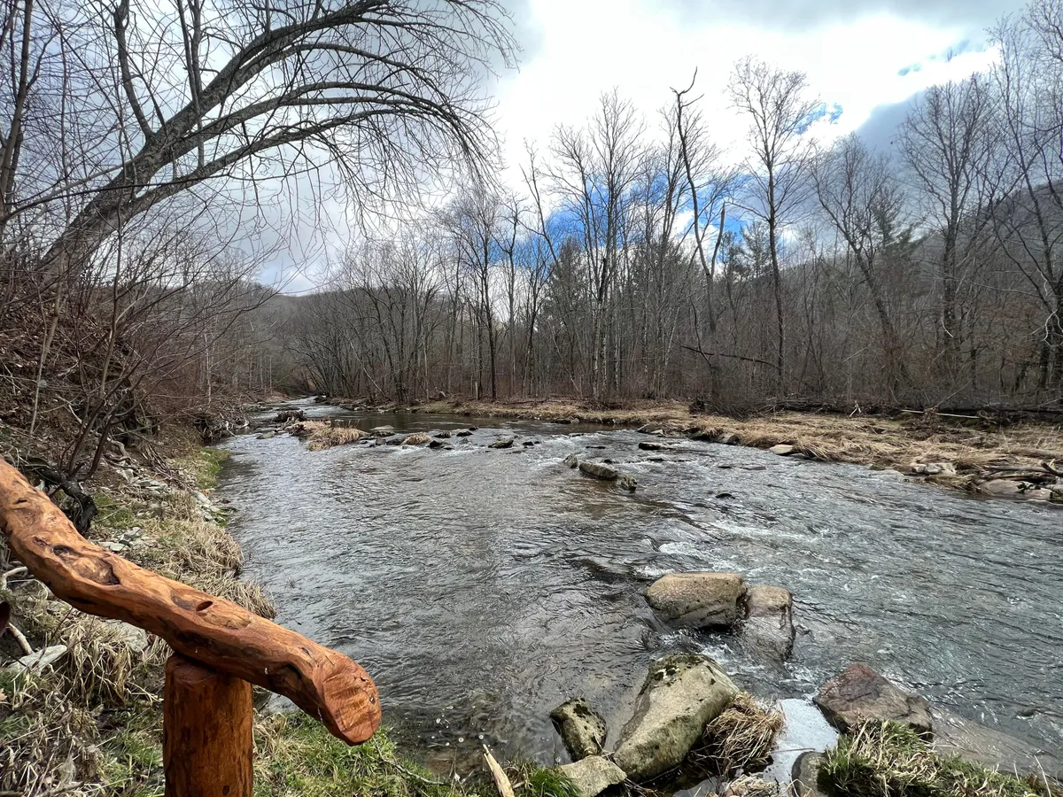 A peaceful riverside view featuring a flowing river bordered by rocky banks and bare trees, framed by a wooden railing. The natural, untouched feel of the setting suggests opportunities for fishing, reflective walks, or simply soaking in the calm of nature.