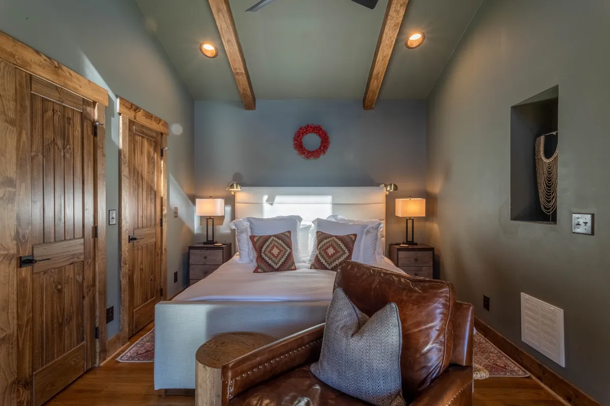A warmly decorated bedroom featuring a bed with plush pillows, wooden accents, a leather armchair, and soft table lamps. There’s a welcoming wreath on the wall and natural light streaming in, creating an inviting space ideal for relaxing, reading, or enjoying quiet moments away from work demands.