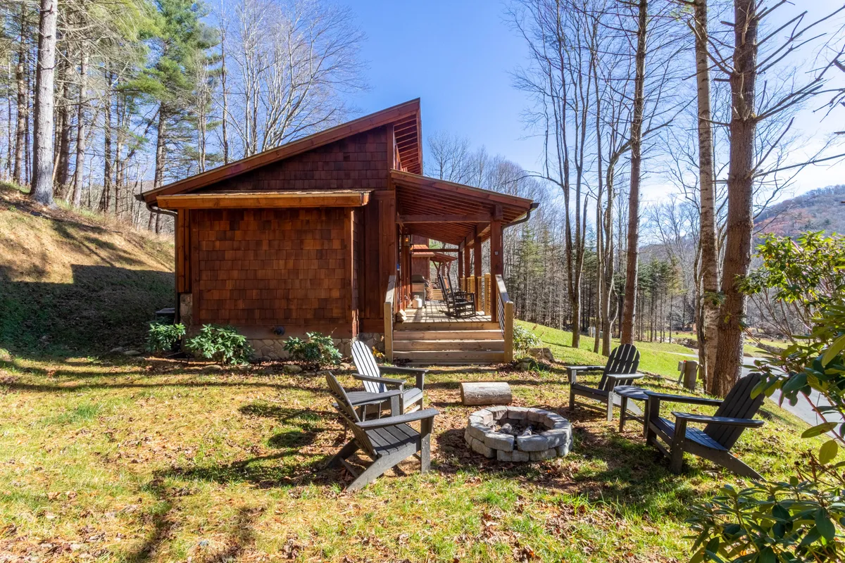 A charming wooden cabin nestled among trees, with a cozy fire pit surrounded by Adirondack chairs. The scene is set for intimate outdoor gatherings, with soft lighting suggesting the magic of dusk or evening. The cabin's inviting porch hints at tranquil mornings and peaceful relaxation, while the natural backdrop emphasizes seclusion and privacy.