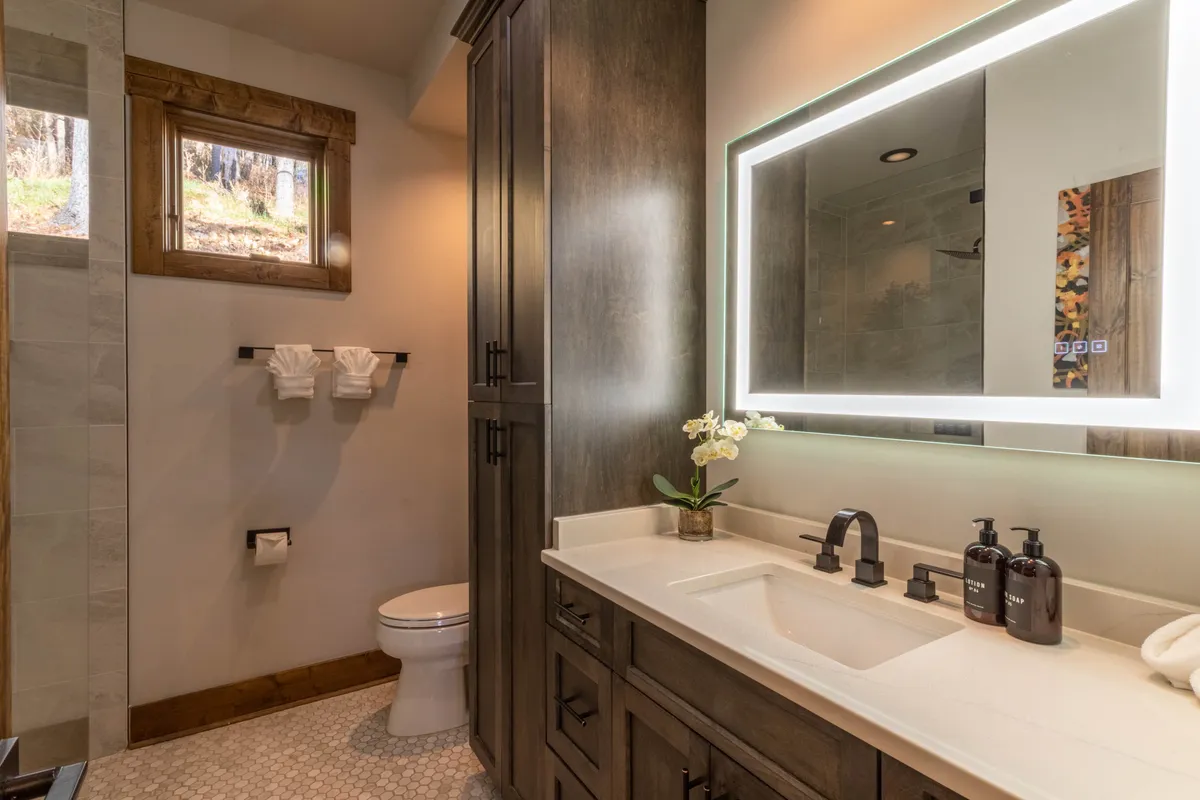This image focuses on a modern bathroom vanity with an LED-lit mirror, sink set in a stone countertop, elegant wood cabinetry, and a window that lets in natural light. The space feels serene, with thoughtful touches inviting relaxation and self-care.
